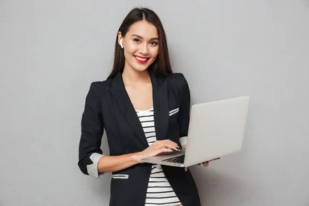 93811359 happy asian business woman holding laptop computer and looking at the camera over gray background